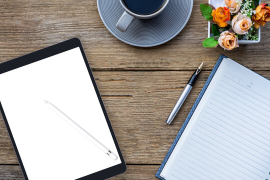 Mockup image of black digital tablet computer pc with blank white screen, diary notebook, cup of coffee and flowers on rustic wooden table background. Top view. Flat lay.