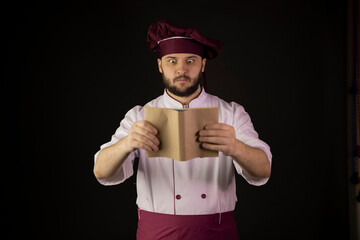 Surprised handsome chef man in uniform holds cookbook. Сook in apron looking on recipe-book on black background. Gastronomy, cooking workshop, delicacy, culinary school, gourmet food concept