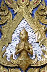 Fototapeta na wymiar Gros plan sur un petit buste de Bouddha doré en position de prière, les mains jointes, sur fond de mosaïque en miroir, et dorures représentant des flammes ou de la végétation sur un mur peint en bleu.