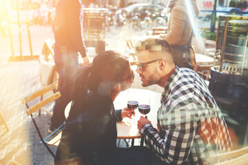 Nice young couple is spending the honeymoon in Barcelona. Boyfriend and girlfriend are sitting in the cozy cafe, holding the wine glasses and looking at each other.  View thought window of coffee shop