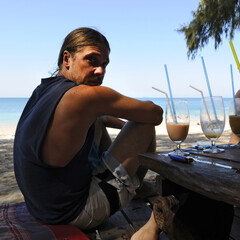 Jeune homme  en vacances assis sur une plage de sable fin au bord de la mer d'Andaman et buvant un cocktail au petit matin.