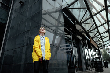 A guy in a yellow jacket and white shirt poses near a gray building with a metal structure