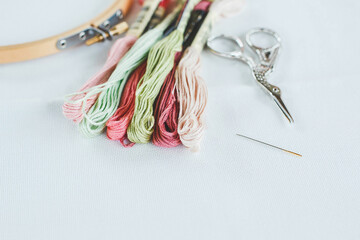 multi-colored cotton floss thread for cross-stitch, a needle, scissors and a wooden hoop on a white...