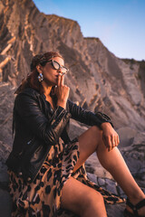 Lifestyle, a young brunette in a leather jacket and leopard dress on a sunset on the coast wearing sunglasses. Young man smoking and looking out to sea at sunset