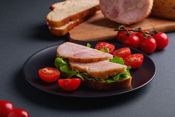 Sandwich with turkey ham meat, green salad and fresh cherry tomatoes slices on black plate near to ingredients on cutting board, grey minimal background, angle view macro