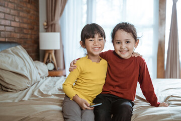 two little girls hugging each other laughing happily looking at the camera while sitting on the bed and holding a smartphone