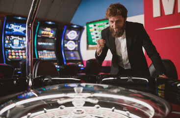young man in a modern black suit stands next to a roulette machine and looks forward to winning at games of chance