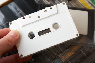 Male fingers hold white audio cassette without markings. Box with tapes in background. Close-up shot