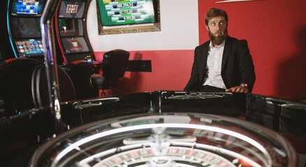 young man in a modern black suit stands next to a roulette machine and waits impatiently for a win