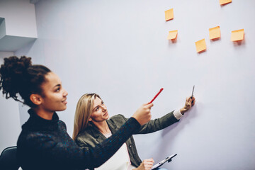 Two female colleagues pointing on stickers with creative ideas on wall discussing strategy for working process, clever woman coach explaining information to african american student during lesson
