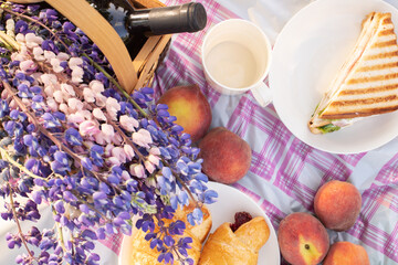 Dinner outdoors in summer. Basket with flowers, wine, peaches, sandwiches on the tablecloth on a Sunny day, top view