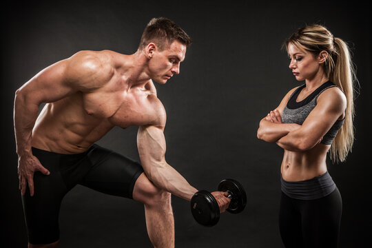 Fitness in gym, sport and healthy lifestyle concept. Couple of athletic man and woman showing their trained bodies on black background. Two bodybuilder models standing and demonstrating tight muscles.
