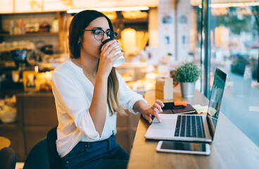 Female student drinking coffee and using laptop in modern workspace