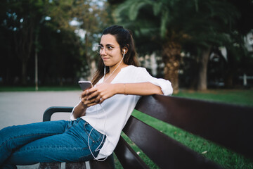 Positive young lady listening to music on bench in park