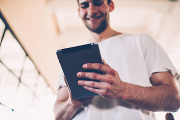 Selective focus on digital tablet in hand of cheerful male making booking via application, smiling hipster guy satisfied with low prices in online retail store making shopping using portable pc