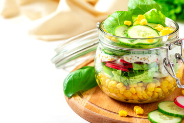 Fresh vegetable salad in a mason jar close-up. Space for text. Salad of corn, radishes, cucumbers and lettuce in a glass jar on white background. Vegetarian food, healthy diet food.