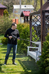 Handsome 40s man washes wooden swings in country house garden with high pressure washer. Spring cleaning concept. modern cleaning technologies.