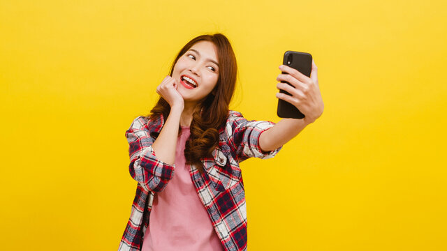 Smiling adorable Asian female making selfie photo on smartphone with positive expression in casual clothing and looking at camera over yellow background. Happy adorable glad woman rejoices success.