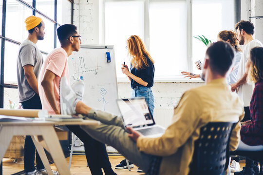 Skilled male coach supervising brainstorming session of trainees presenting their ideas using whiteboard, designers colleagues cooperating  in friendly atmosphere using teambuilding concept in job