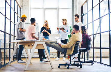 Crew of creative marketing experts presenting to experienced project manager of company sitting with laptop computer  ideas for advertising campaign using flip chart on informal conference meeting