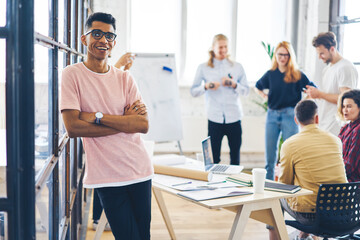 Positive talented afro american male in eyeglasses posing for camera and laughing while team members discussing creative ideas for business startup project during brainstorming session in office