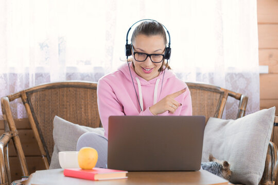 Pretty smiley brunette girl with long dark hair dressed in pink hoodie wearing glasses works remotely with laptop computer at her country house. distance work concept. Online conference concept