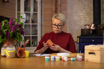 attractive middle-aged woman coming up with drawing for a flowerpot in cozy country house with fireplace Concentrated woman with blond hair dressed casually painting