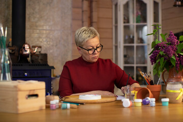 attractive middle-aged woman draws in cozy country house with fireplace Concentrated woman with blond hair dressed casually painting