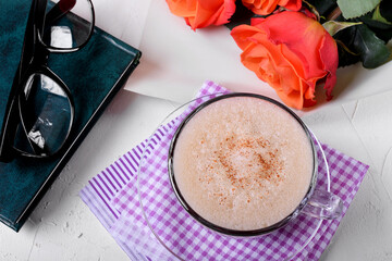 Coffee with foam and cinnamon, book, glasses and roses on the white table