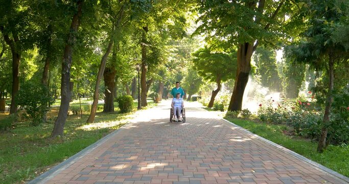 Young nurse spending sunny day with senior woman in wheelchair walking in park. 4k