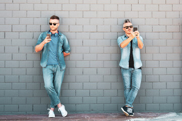 Two young man looking at mobile phone lean against brick wall - Couple of cool guys using smartphone standing outdoor in the street