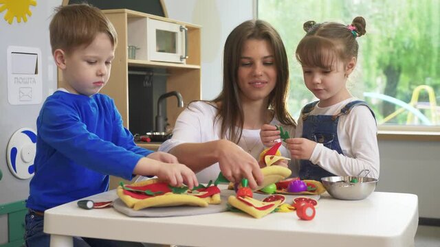 Caucasian girl and boy kid playing and learning at preschool with female teacher. Mother, daughter adn son playing cooking