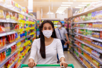 Asian woman wear face mask push shopping cart in supermarket department store. Girl looking grocery to buy  something. During coronavirus crisis or covid19 outbreak. Women wearing protective face mask