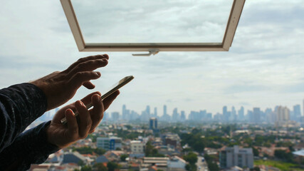 A man types a message on a smartphone against the background of an open window and a panorama of the city of Jakarta. Hands close up.