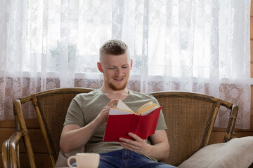 young smiling man reading book with red cover on wicker bench in countryside wooden house against background of basket of yellow apples indoors relaxation concept Free time concept