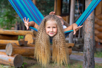 Little happy girl with long blond curly hair sways arms outstretched on a blue-green hammock. Freedom of movement, lifestyle. School holidays, vacation, day off. Summer, carefree, fun. Outdoors.