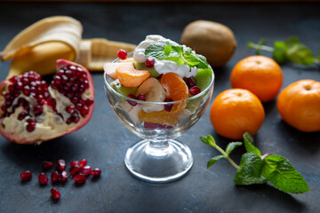 Fruit salad with banana, mandarin, kiwi, pomegranate seeds and yogurt in a glass bowl on a dark background