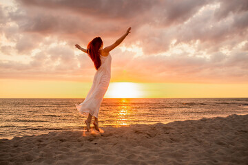 Fototapeta na wymiar Happy young woman in a long white dress looking at the sunset on empty sand beach with her hands up. Freedoom, vacation, meditation