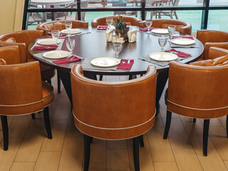 A round table is set for a large company. Soft leather armchairs stand next to a large window. Cafe interior