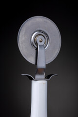 Detail of traditional Italian design pizza slicer with white plastic handle and stainless steel round slice head against a dark background. Studio low key object still life.