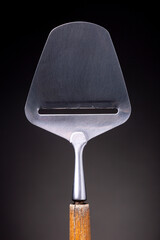 Closeup of traditional Dutch design cheese slicer [kaasschaaf] with wooden handle and stainless steel slice head against a dark background. Studio low key object still life.