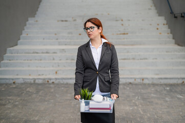 An upset unemployed woman is standing with a box of personal items against a gray wall. Business...