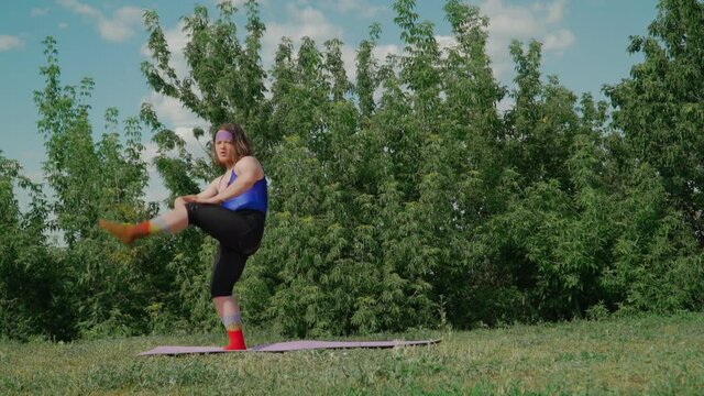 Funny Fat Man with Long Hair in a Tight Female T-shirt Is Engaged in Martial Arts on fitness mat on Green Lawn in City Park. Playful Guy Overweight Depicting a Girl. Sport humor concept