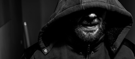 Portrait of an evil bearded man in a hoodie in the dark. Male criminal hides his eyes under the hood.