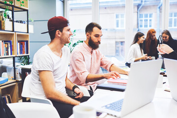 Group of stylish students preparing for exam in library.Hipster freelancer tells joyful story.Business people feel relaxed except male focused on what he is typing.Modern office makes atmosphere light