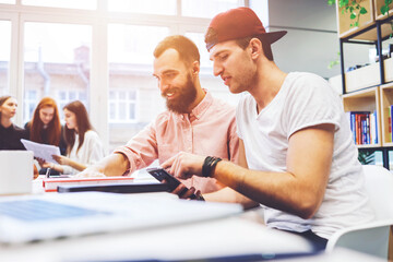 Students in a library are preparing for their report finding the information via the internet using smartphone.Male journalists are reading  funny post on some blog and smile while writing an article