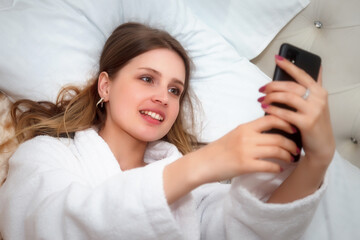 Obraz na płótnie Canvas Girl of Slavic appearance in white coat with phone on bed in hotel room. Pretty female shows acting emotions. Young charming woman is sitting on sofa in bedroom and playing with her feelings. Relax