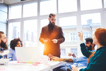 Male talented teacher answering question of students during lecture about innovating technologies and their role in mass media showing modern digital devices using free wireless internet in classroom