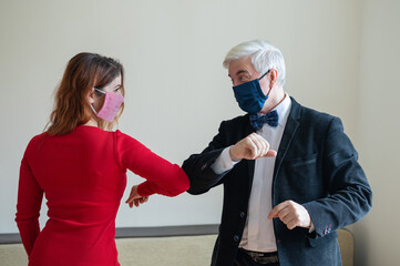 A mature gray-haired man in a suit and a red-haired woman in a red dress wearing in medical masks maintain a social distance and greet their elbows. An alternative to shaking hands and hugs.