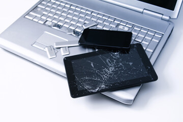 A silver laptop with a broken keyboard, tablet with a cracked display and black phone. A close-up picture of part of broken laptop and cracked screen on a tablet.